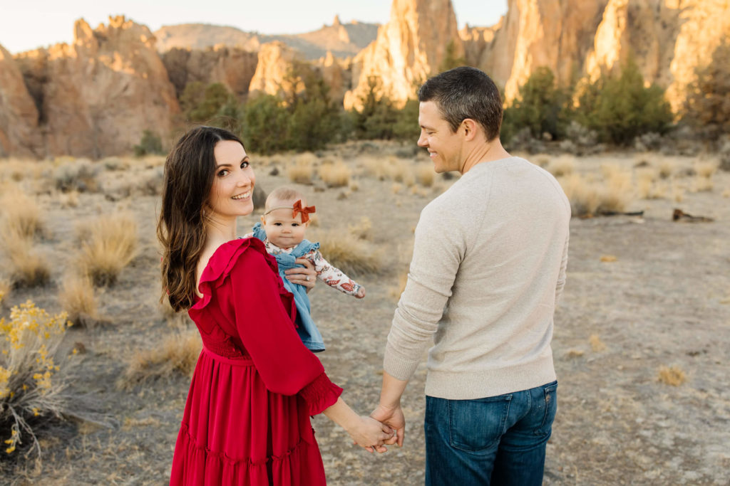 woman in red dress looking over her shoulder while holding her baby and husbands hand