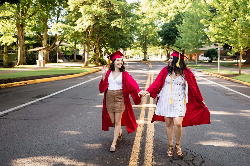 two girls wearing red graduation gowns walking down street holding hands