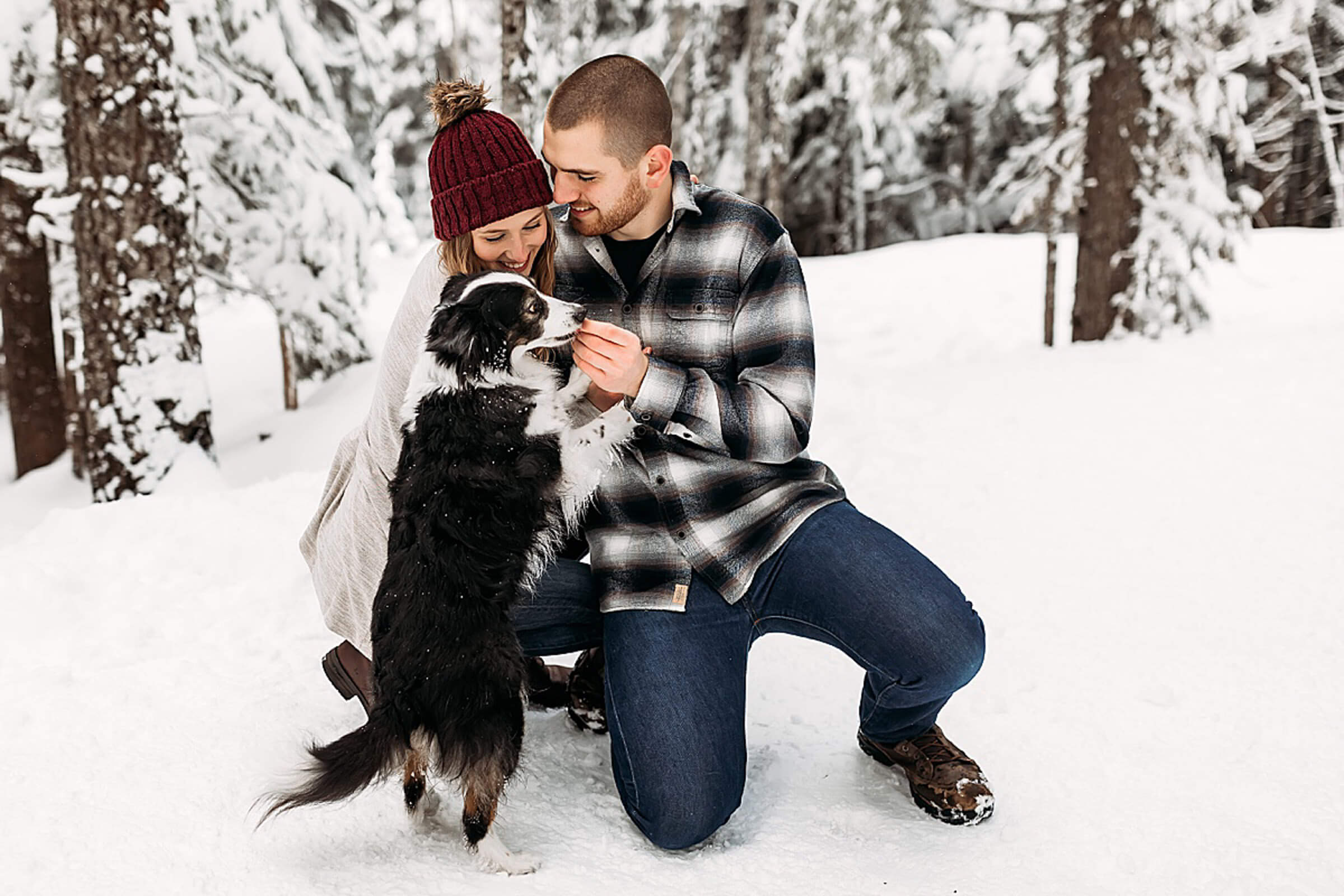 male and female kneeling in snow while their puppy jumps up