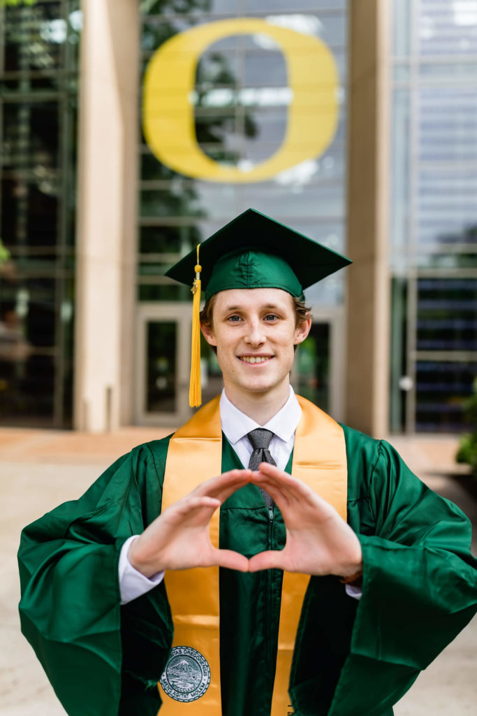 boy in University of Oregon graduation cap and gown in front of lillis complex
