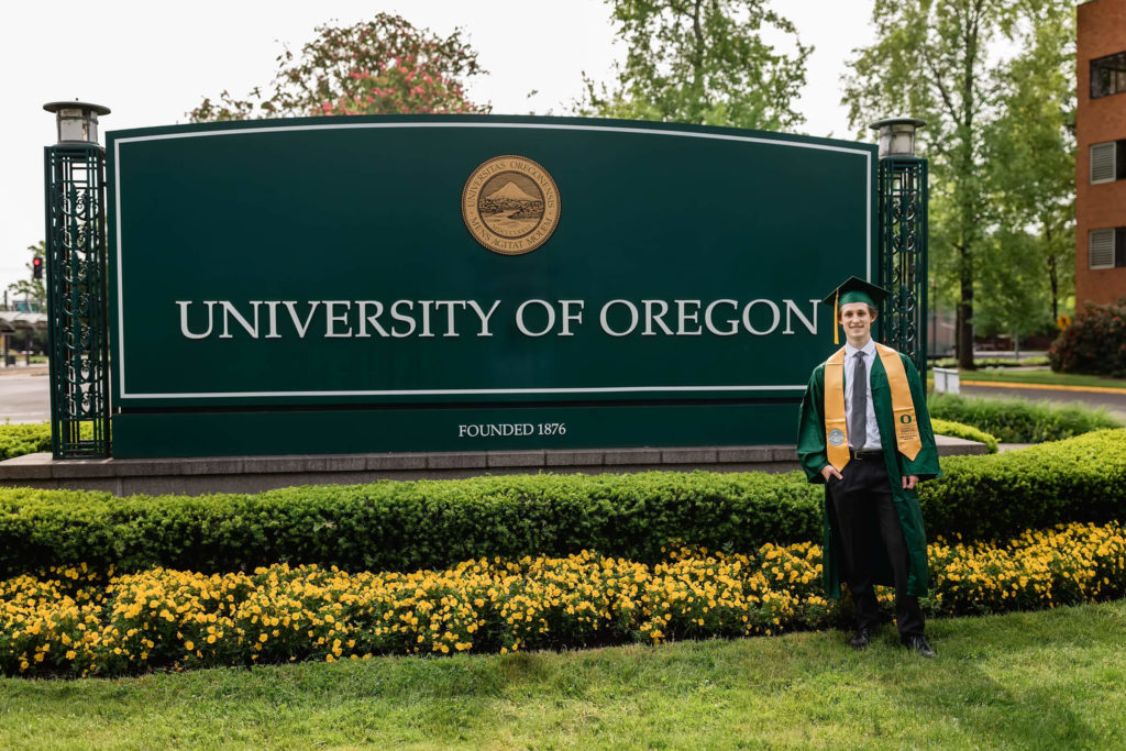 male standing in university of Oregon graduation cap and gown in front of UO sign