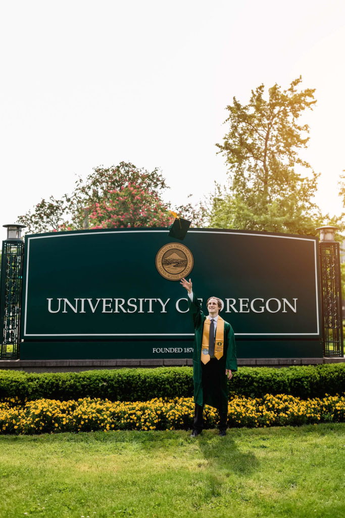 male standing in front of university of Oregon sign throwing his graduation cap in the air