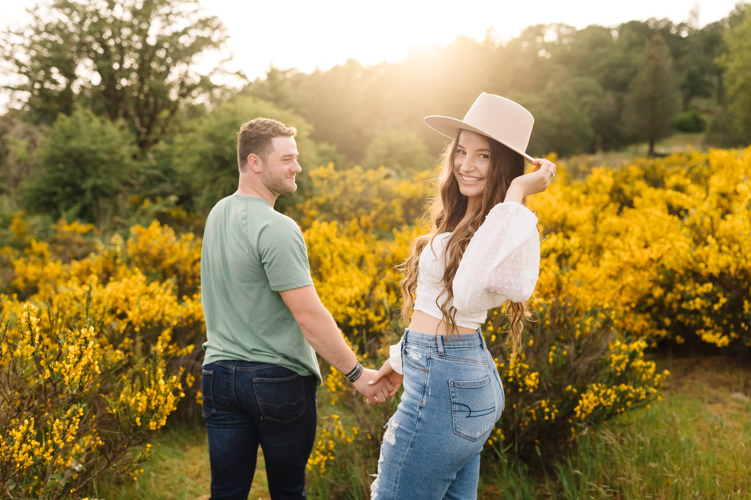 man in green shirt leading girl in white top through a field of yellow flowers during engagement photos