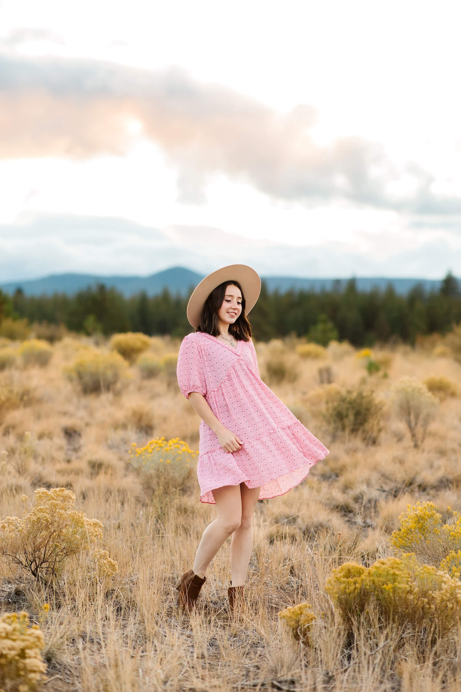 brunette girl in pink dress and tan rancher hat standing in open field in bend