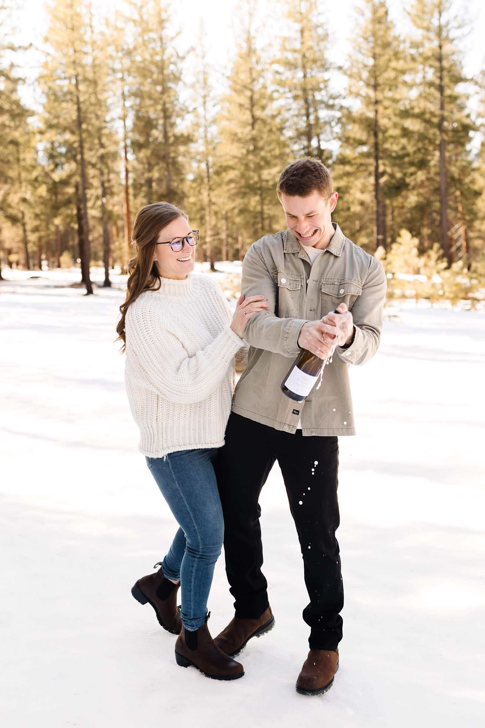 man and woman in the snow shaking bottle of champagne during engagement shoot