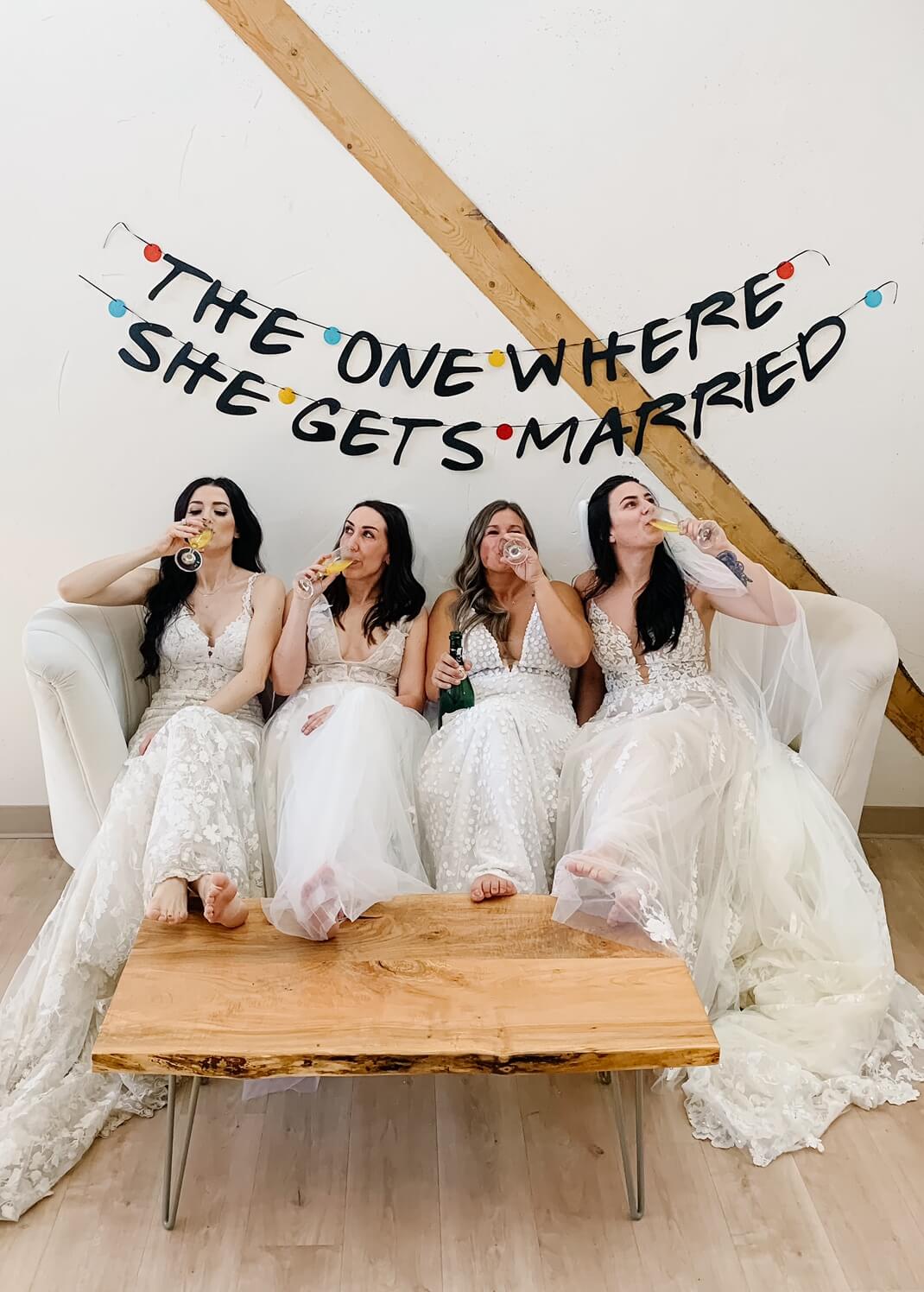 four girls sitting in wedding dresses on a couch drinking champagne at bespoke bride