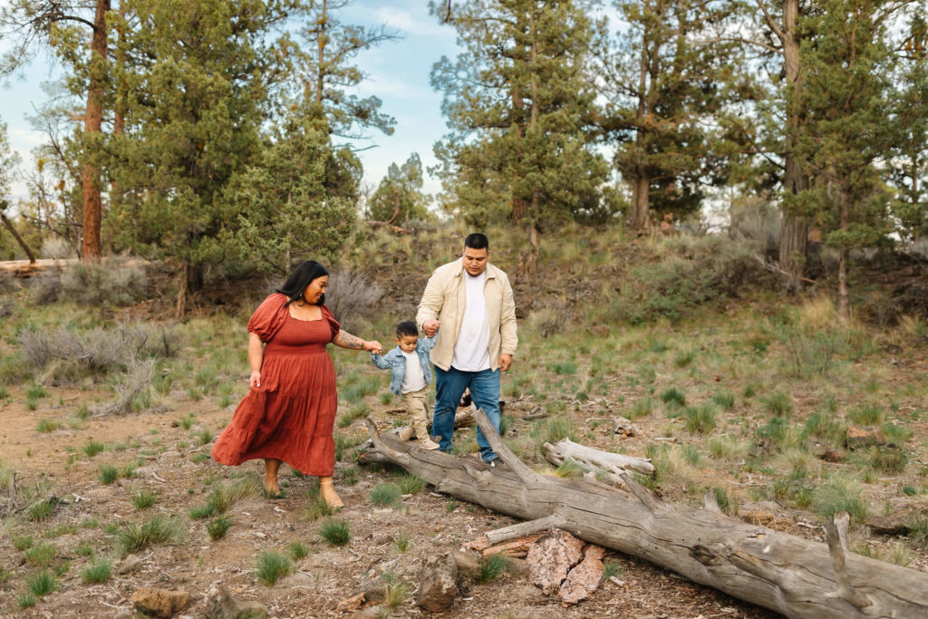 mom in red dress and dad holding hands with little boy in denim jacket walking across log