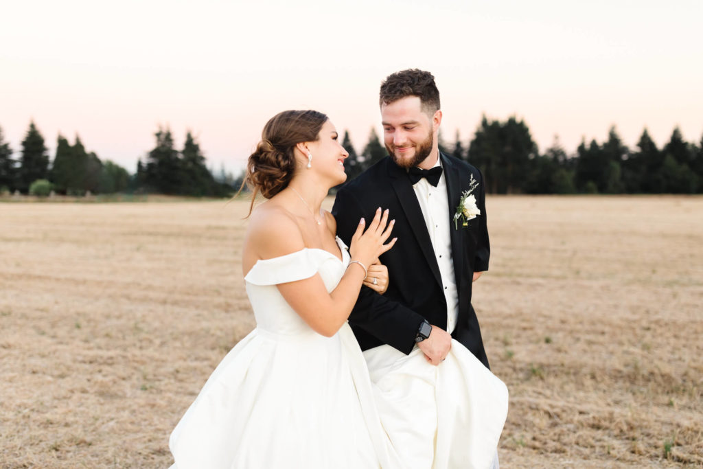 girl in white off the shoulder ball gown wedding dress and groom in black tuxedo running through grassy field