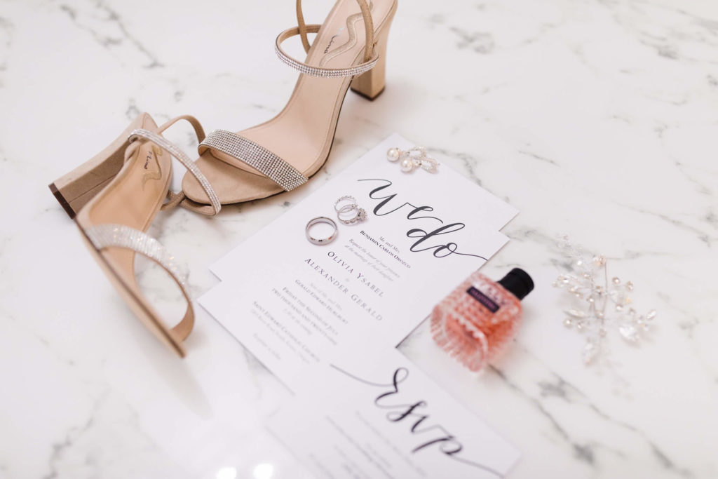 tan wedding heels laying on white marble table with pink perfume bottle and wedding invitations