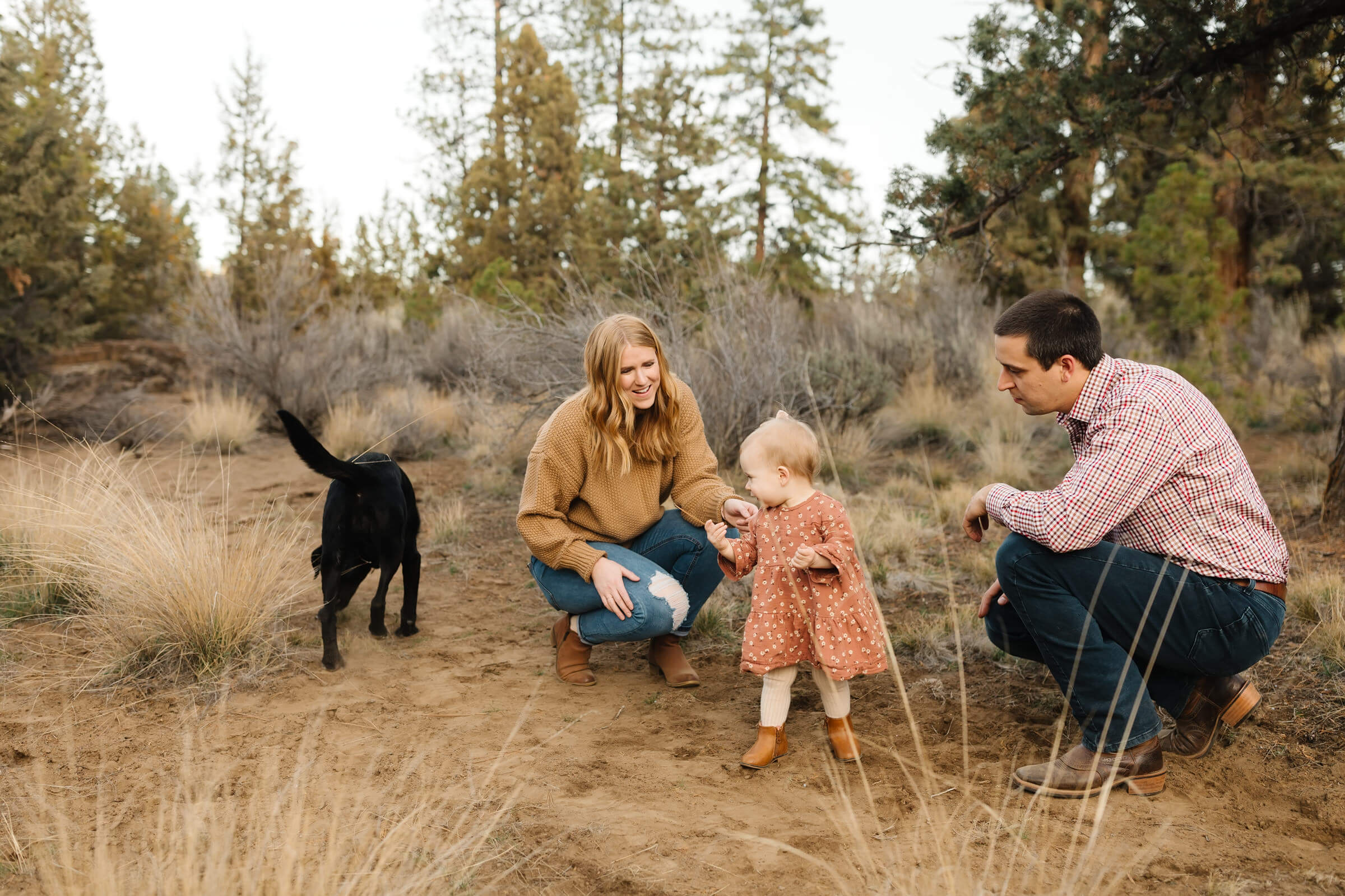 man and woman sitting on a blanket in the dirt with young blonde daughter and black lab