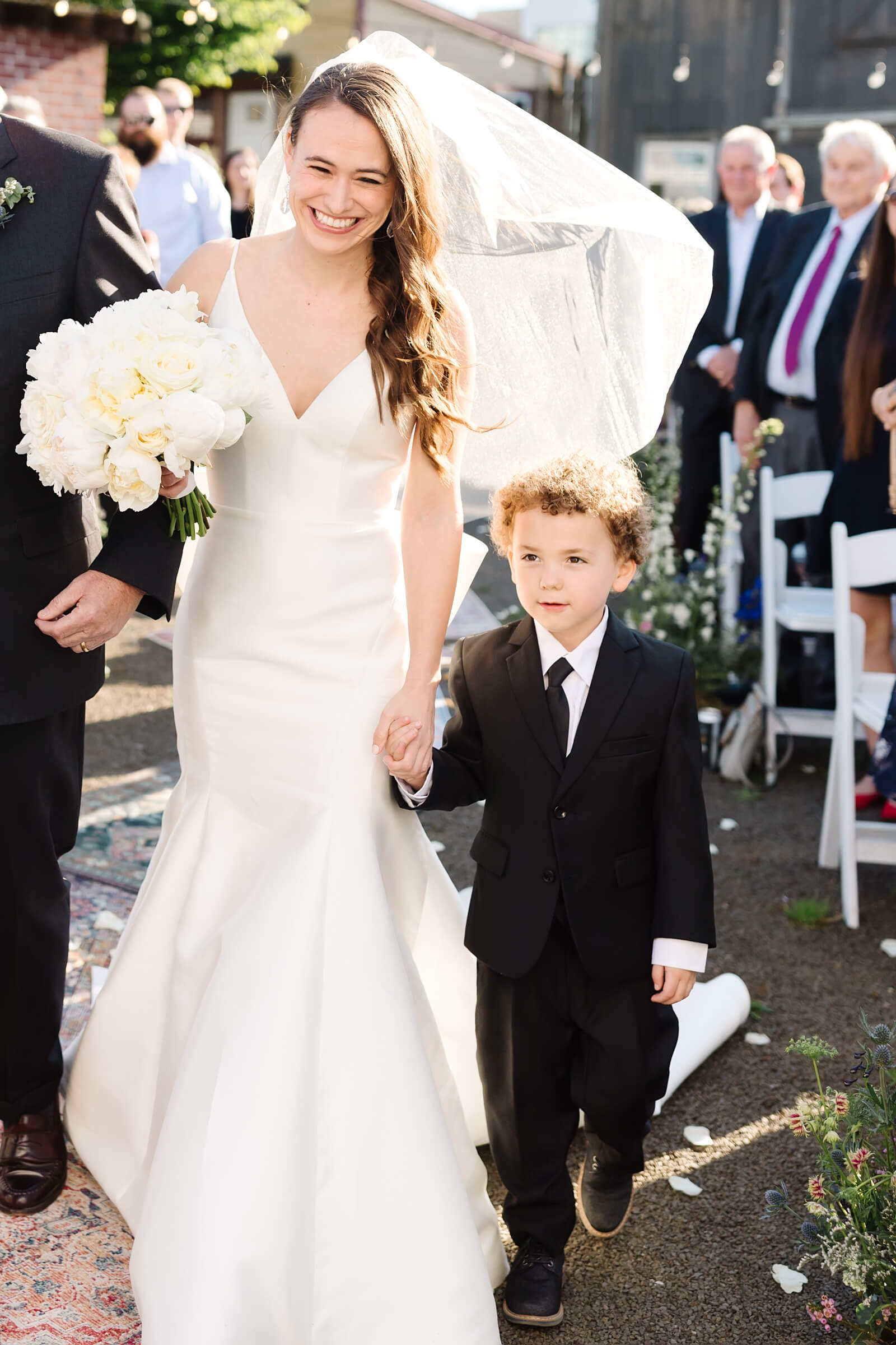 little boy in black suit holding hands with bride walking down aisle