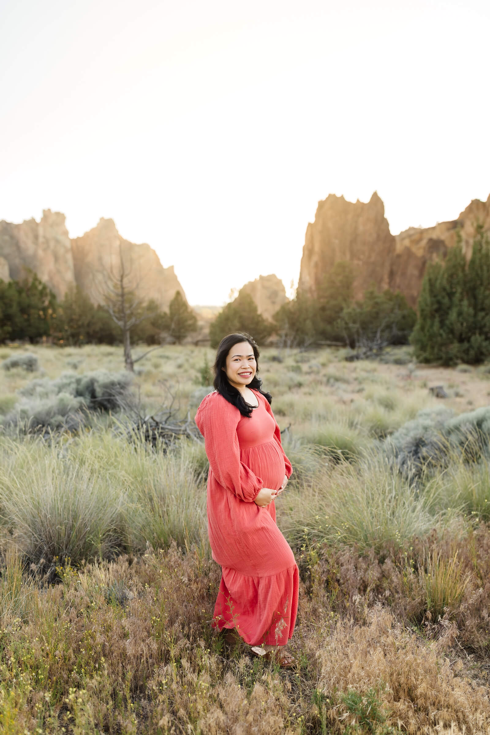 woman with black hair wearing coral dress holding pregnant belly during smith rock maternity photoshoot