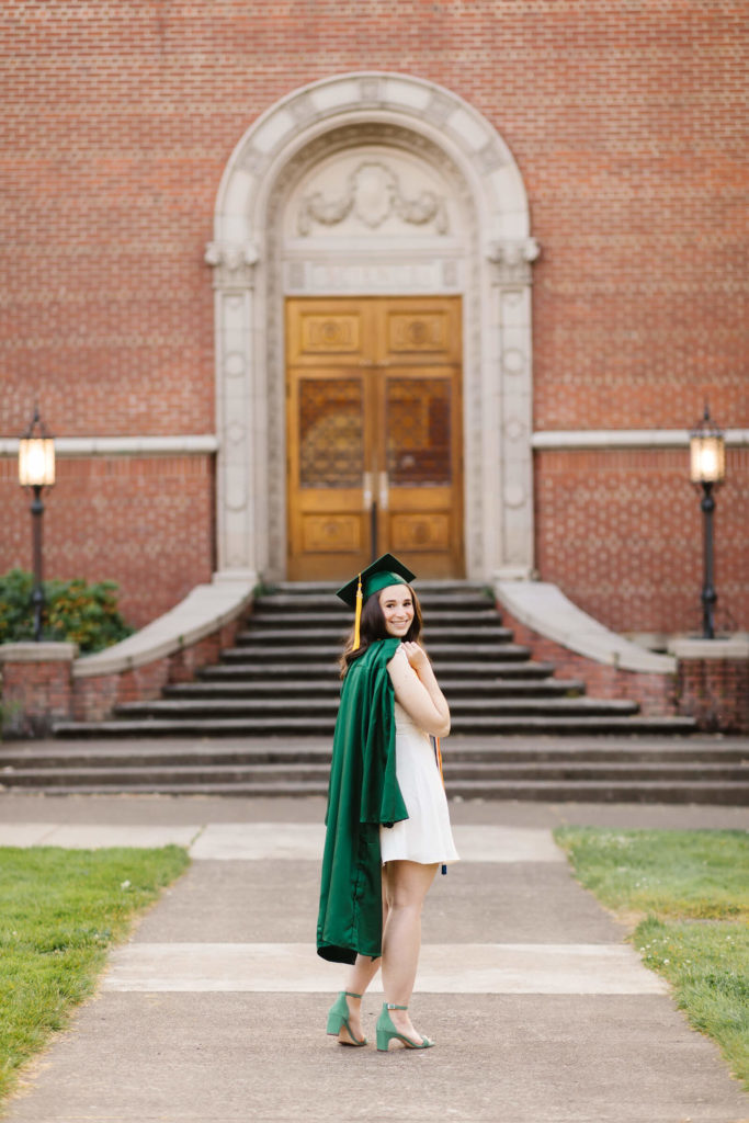 University of Oregon student with green graduation gown slung over shoulder stands in front of brick building