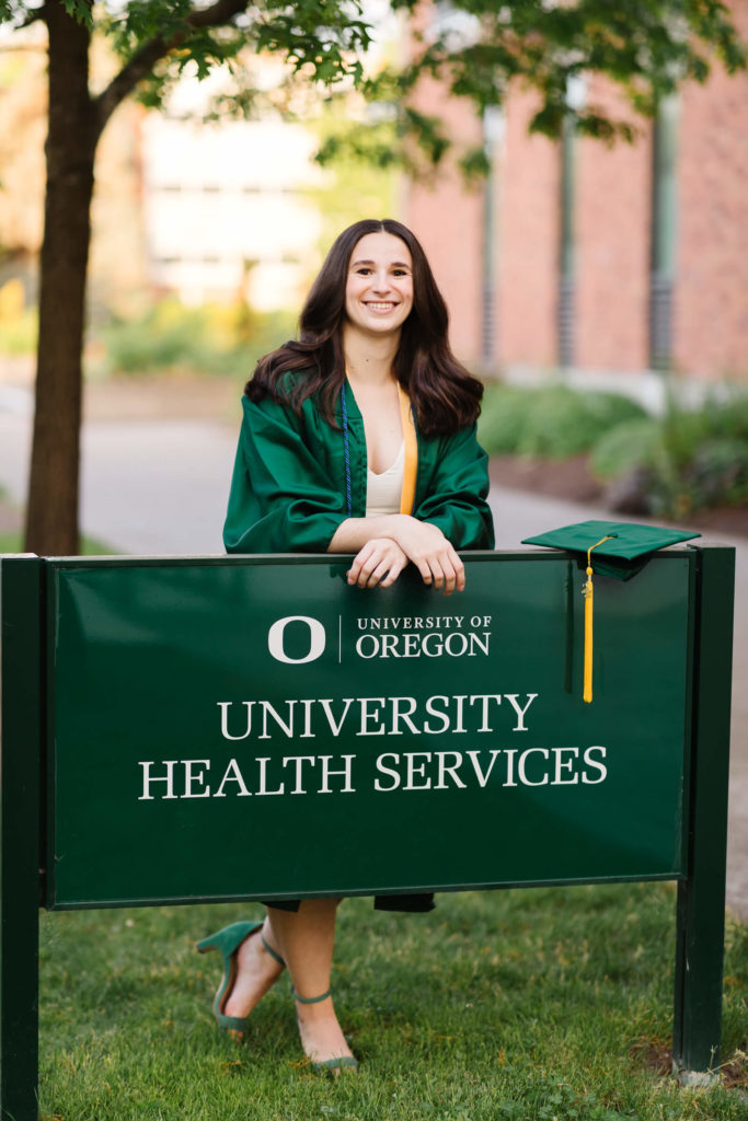 brunette in green UO graduation gown stands in behind university of Oregon health services sign 