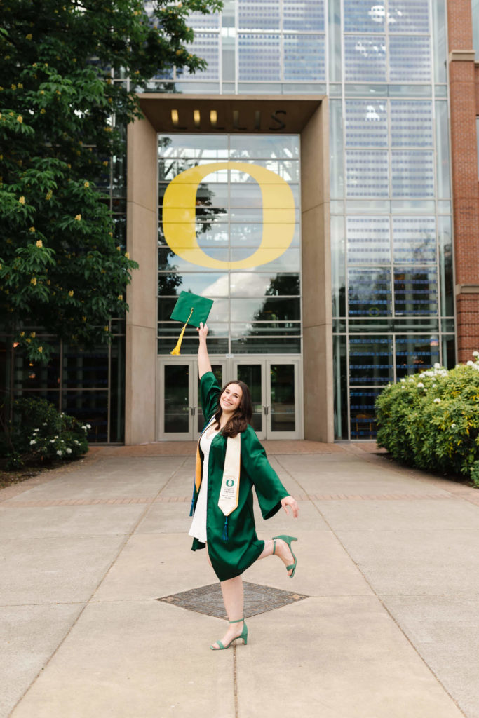 brunette female in green graduation gown tossing graduation cap in front of lillis complex at UO