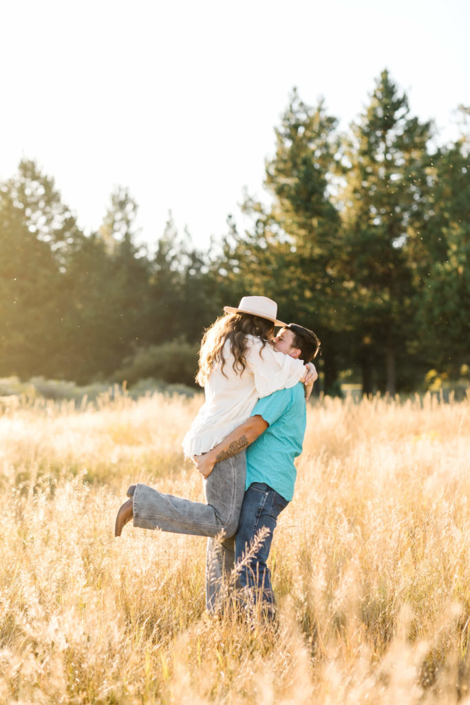 brunette male in teal button up lifting girl in white while in golden hay field