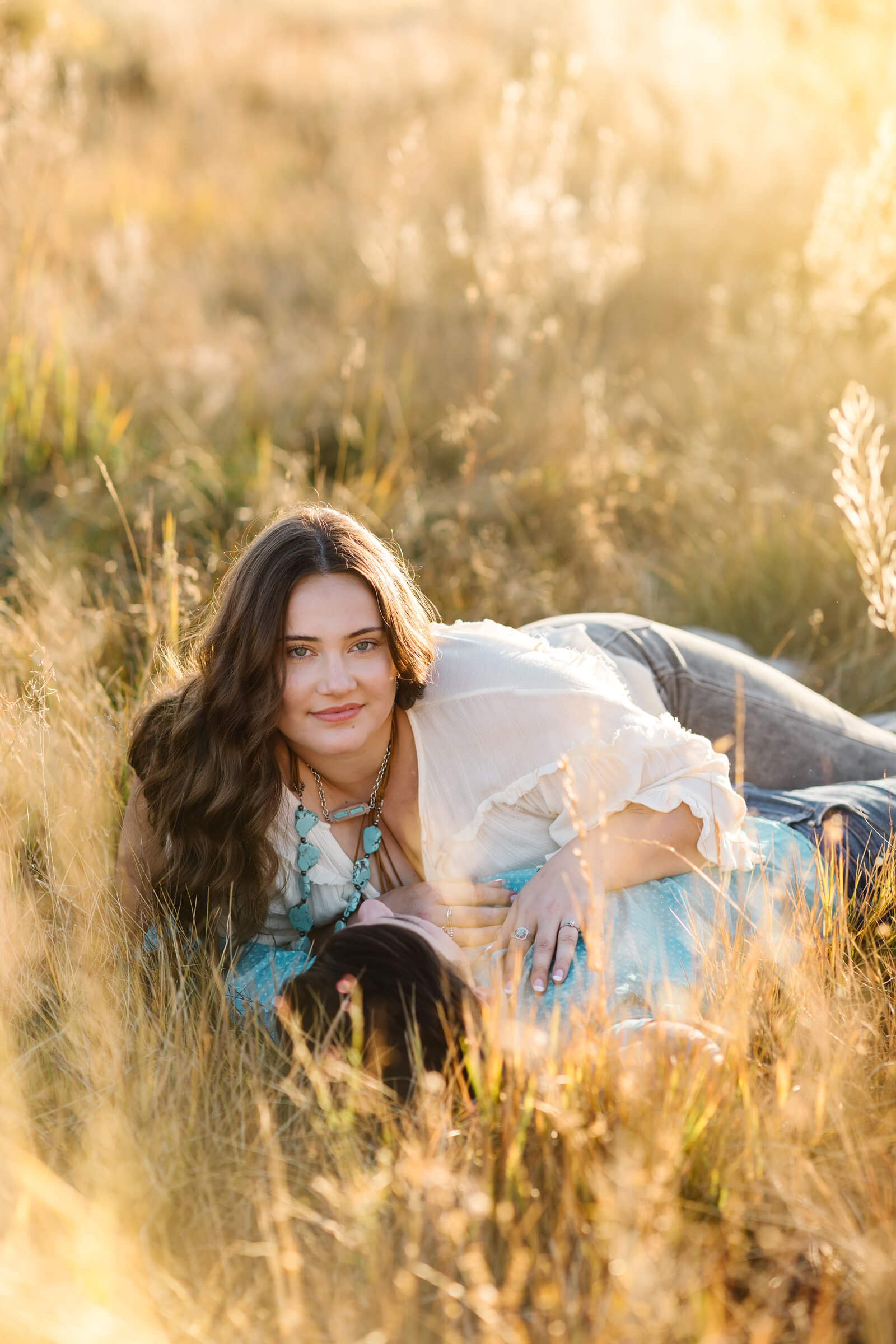 brunette girl in white laying with man in teal button up in golden hay field looks at camera