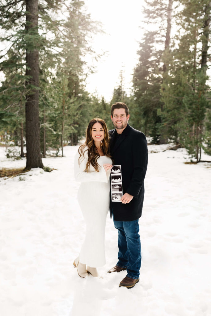 man and woman smiling and standing in the snow holding sonogram during pregnancy announcement shoot