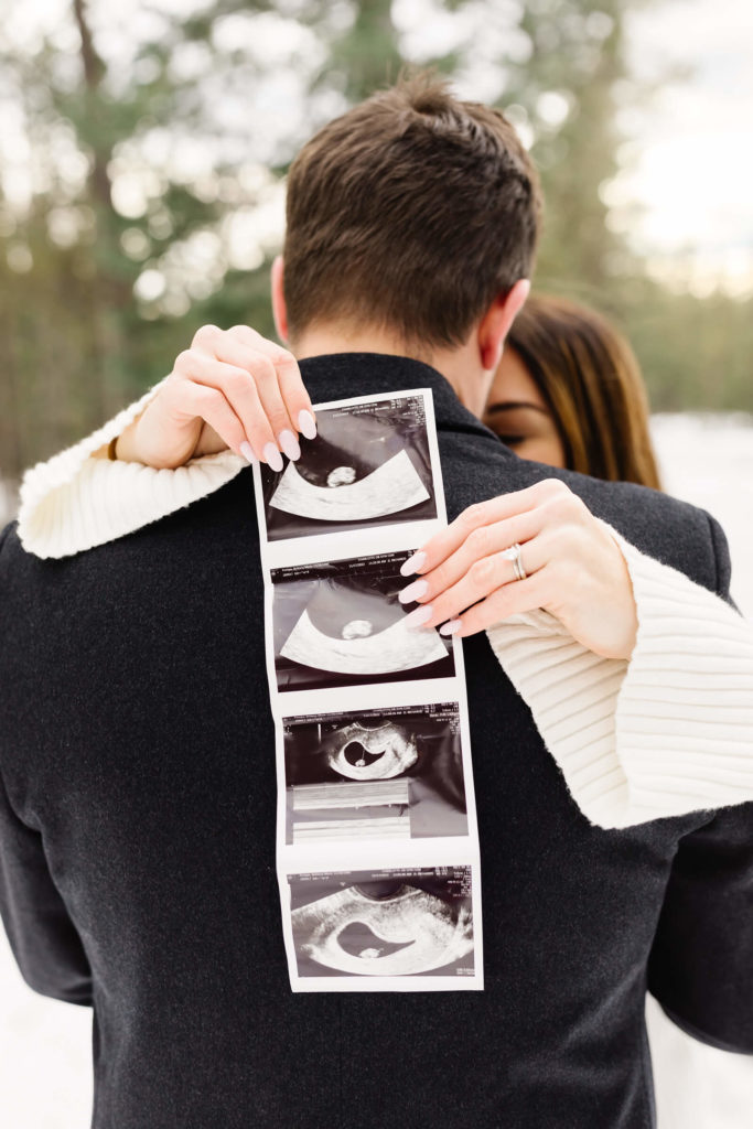 woman holding sonogram behind husbands back while hugging him during pregnancy announcement shoot