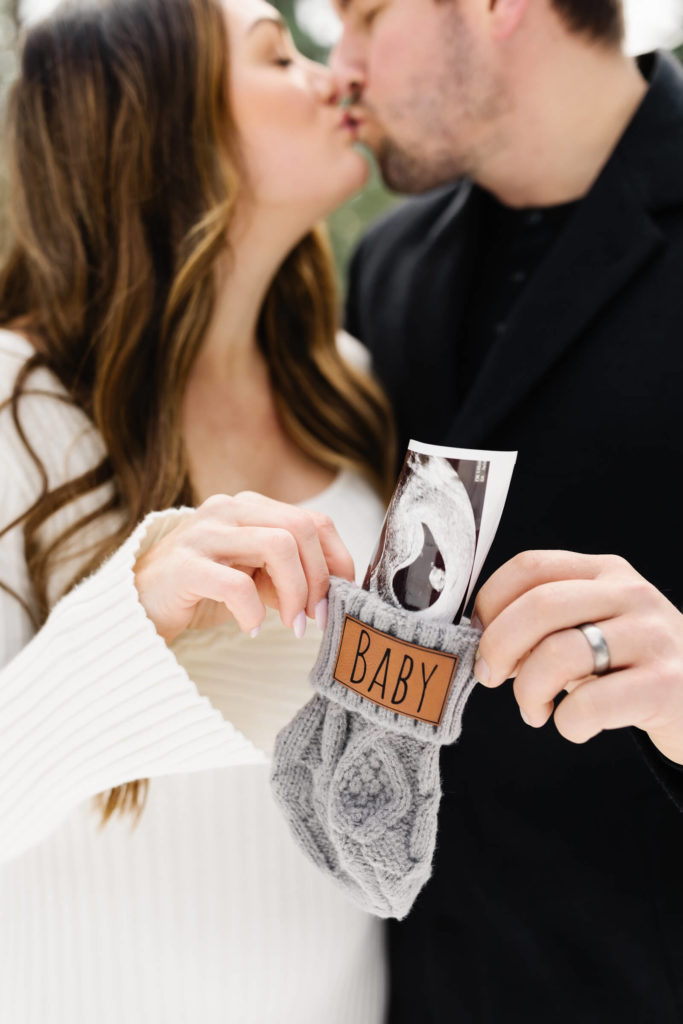 man and woman kissing while holding a tiny stocking that says baby with sonogram rolled up inside