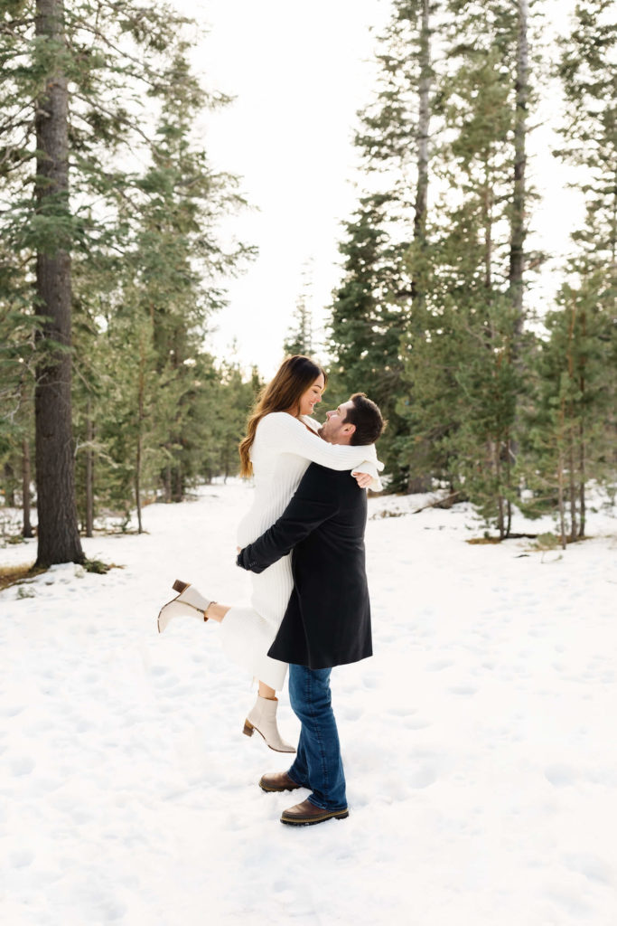 man lifting woman in white knit dress in the snow during pregnancy announcement shoot