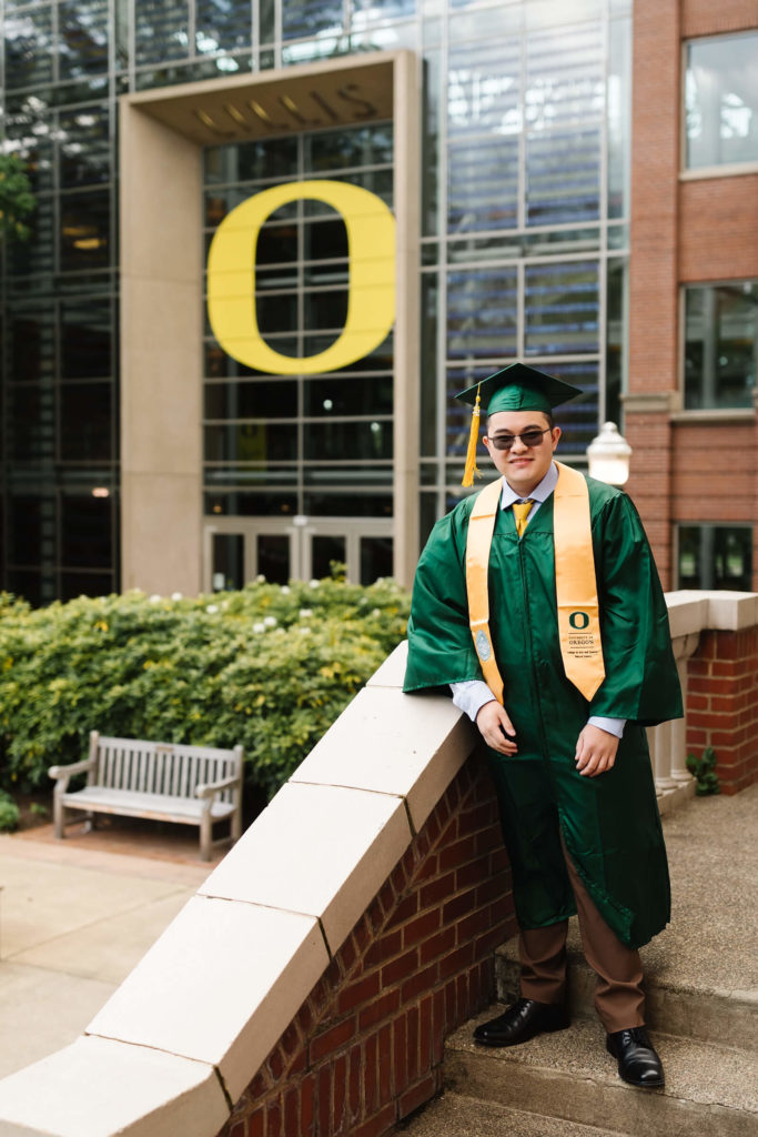 male student in university of Oregon green graduation gown outside Lillis business complex smiling during Hayward field graduation photos