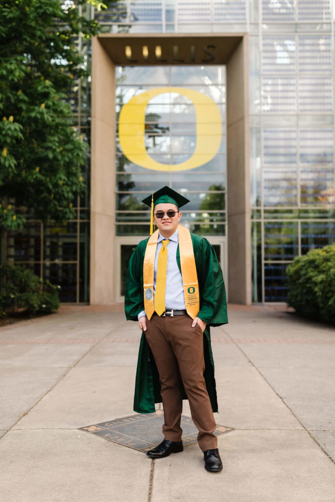 male student in university of Oregon green graduation gown outside Lillis business complex smiling at camera during Hayward field graduation photos