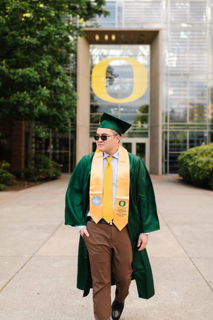male student in university of Oregon green graduation gown outside Lillis business complex walking towards camera during Hayward field graduation photos