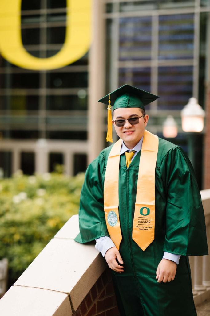 male student in university of Oregon green graduation gown outside Lillis business complex smiling during Hayward field graduation photos