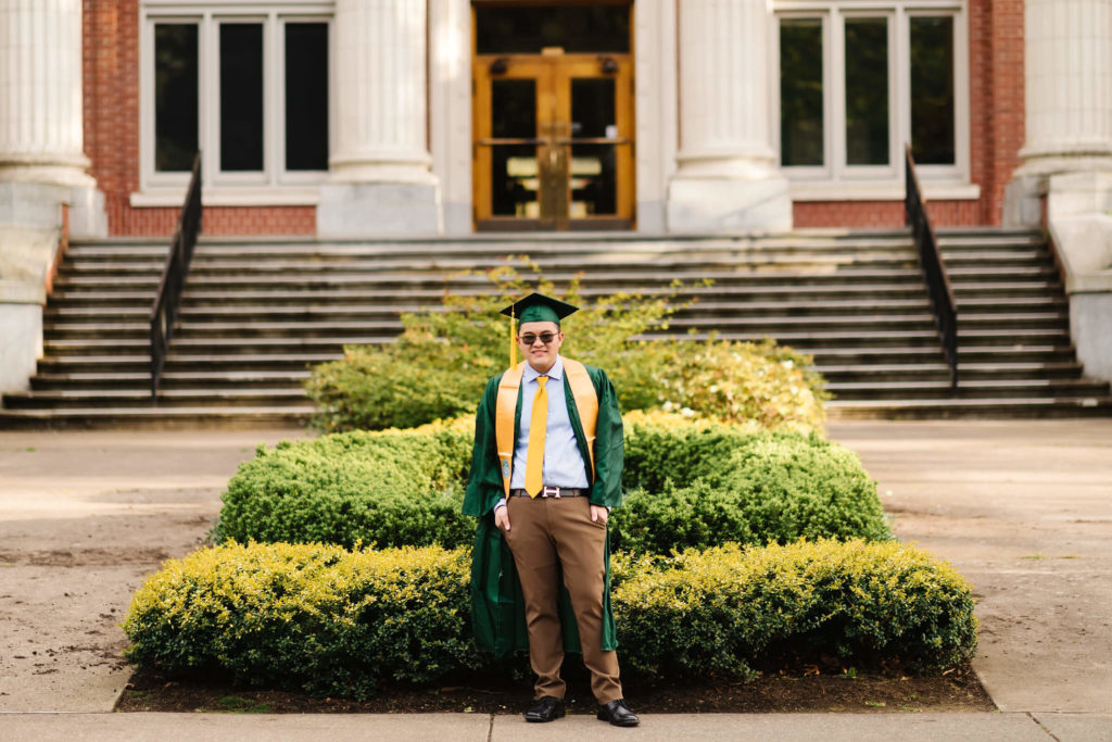 University of Oregon male student standing in front of Johnson Hall and holding yellow graduation stoll during Hayward field graduation photos