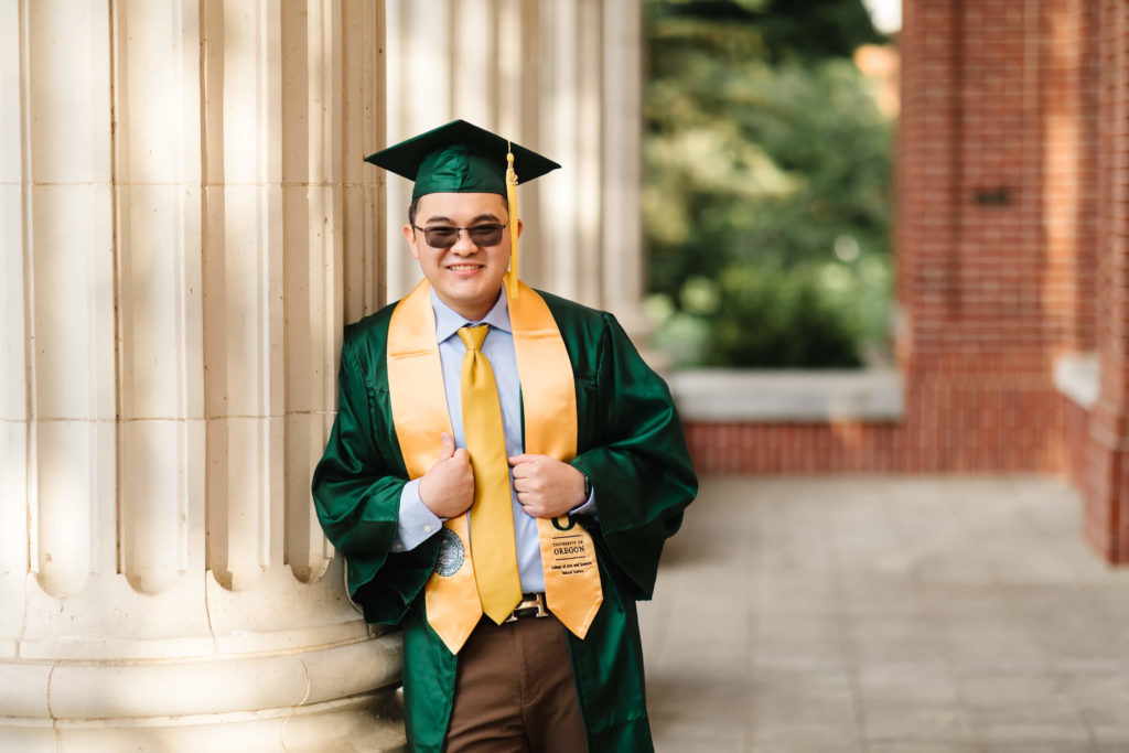 University of Oregon male student leaning against white columns at Johnson Hall and holding yellow graduation stoll during Hayward field graduation photos