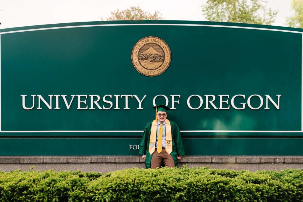 male U of O student sitting in front of University of Oregon sign during hayward field graduation photos