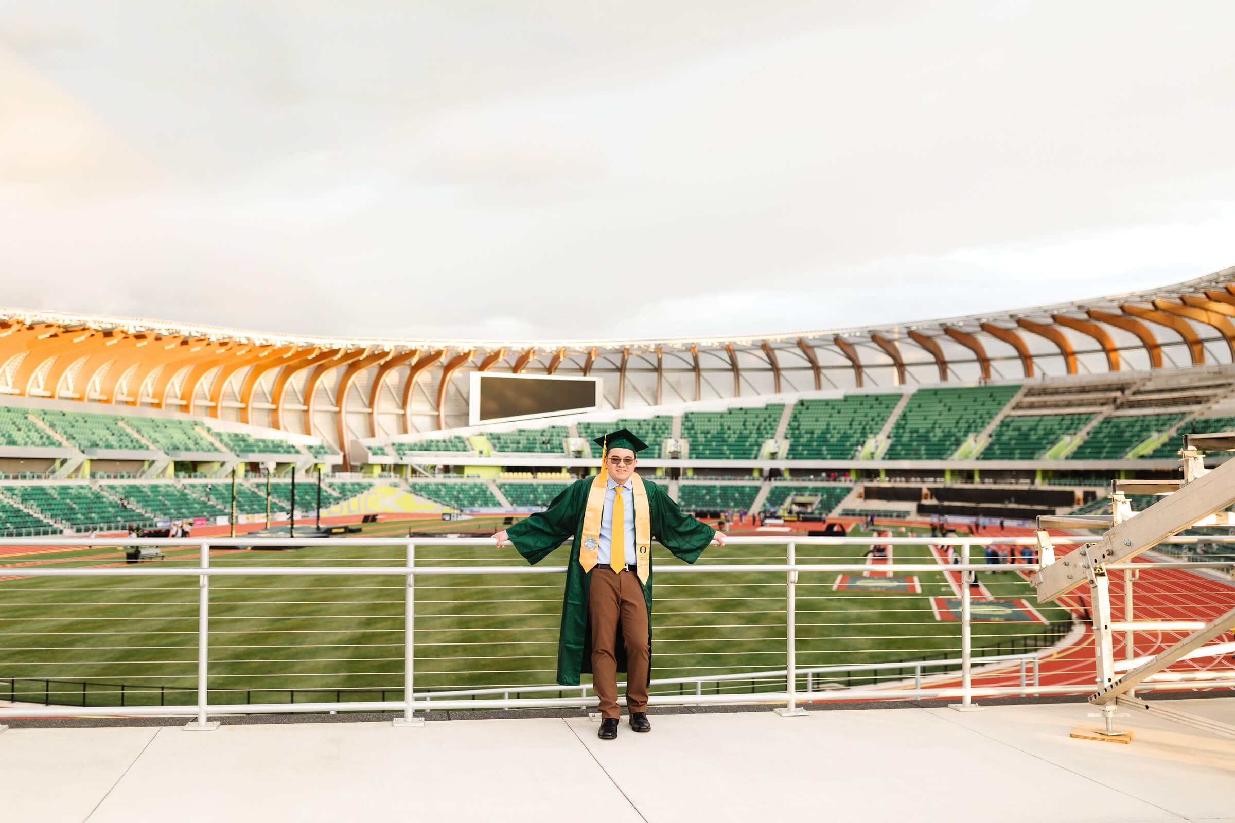 male university of oregon graduate standing in green graduation cap + gown in front of Hayward Field during graduation photos