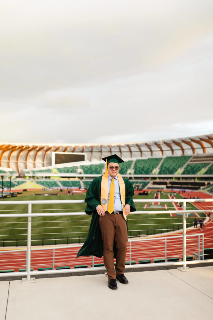 male university of Oregon graduate standing in green graduation cap + gown in front of Hayward Field during graduation photos