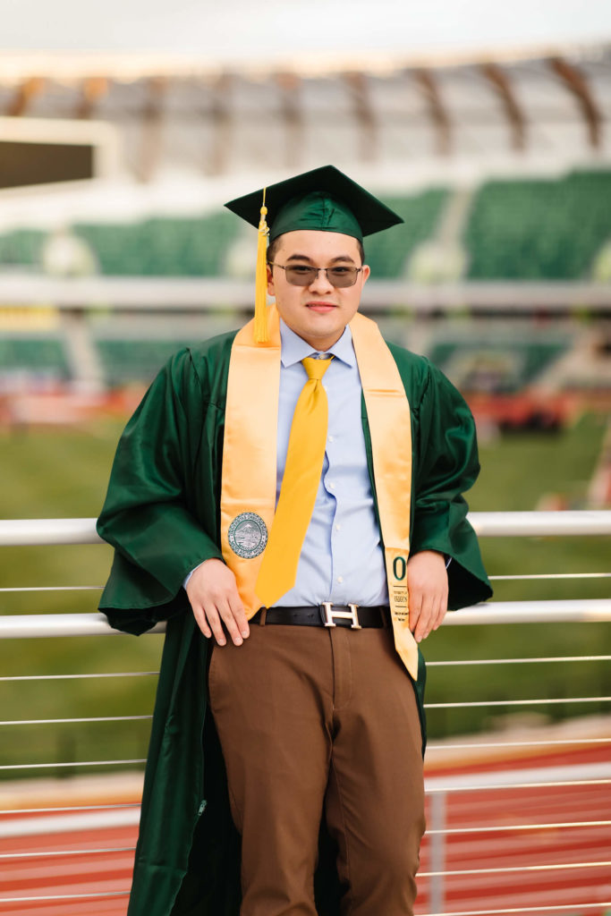 male university of Oregon student leaning of aluminum hand railing in green graduation gown during hayward field graduation photos