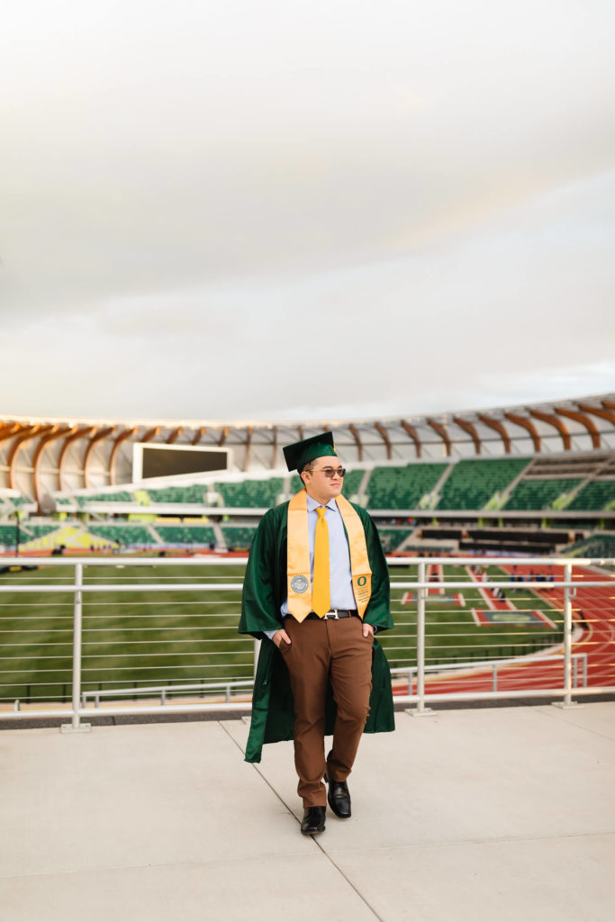 male university of Oregon graduate walking in green graduation cap + gown in front of Hayward Field during graduation photos