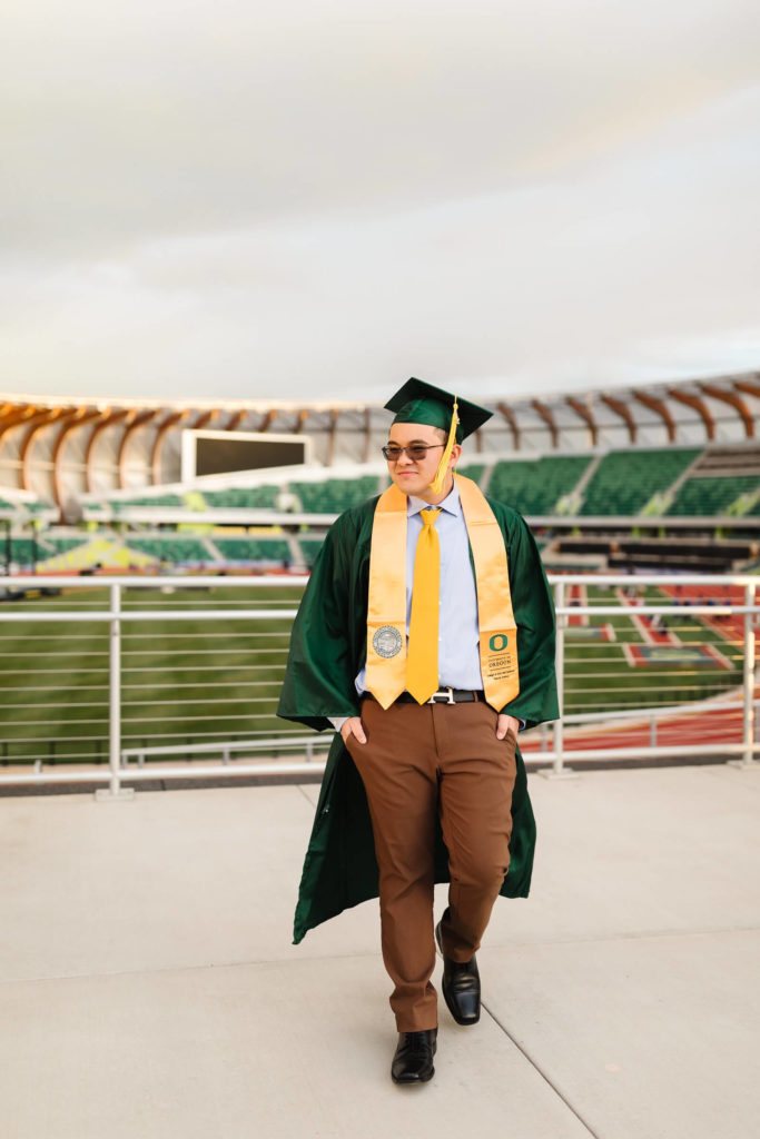 asian male university of Oregon graduate walking in green graduation cap + gown in front of Hayward Field during graduation photos