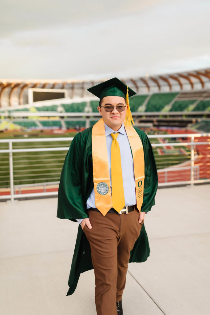 male university of Oregon graduate walking in green graduation cap + gown in front of Hayward Field during graduation photos
