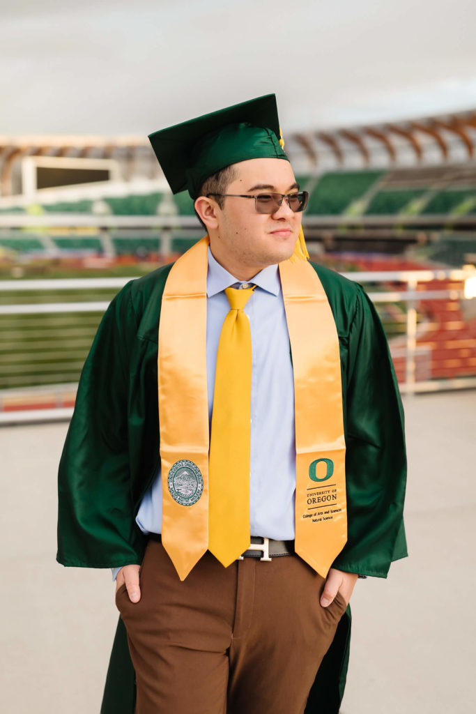 male university of Oregon graduate looking into distance wearing green graduation cap + gown in front of Hayward Field during graduation photos