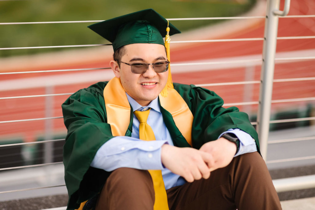 male university of Oregon graduate wearing green graduation cap + gown sitting in front of fence looking over Hayward Field during graduation photos