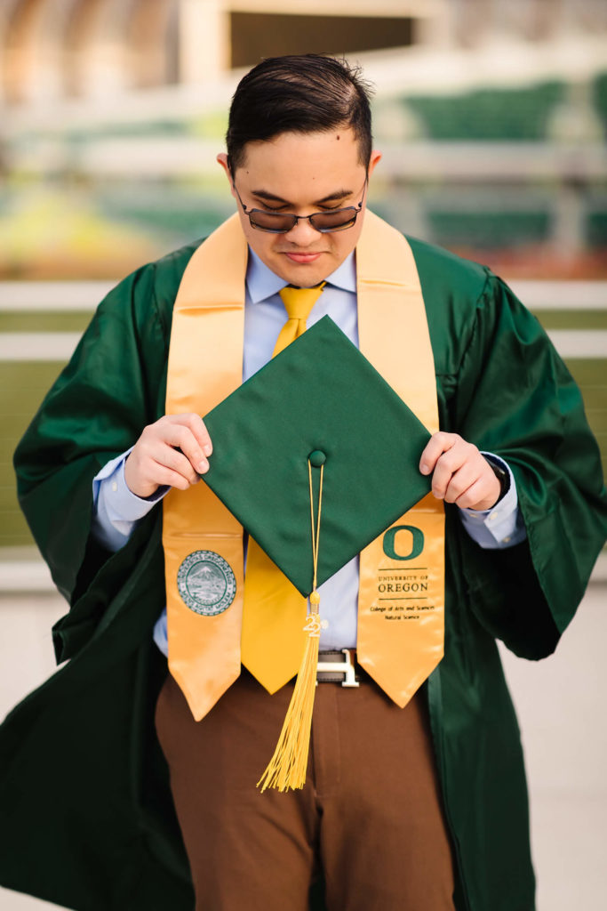 male university of Oregon graduate looking down at green graduation cap standing in front of fence looking over Hayward Field during graduation photos