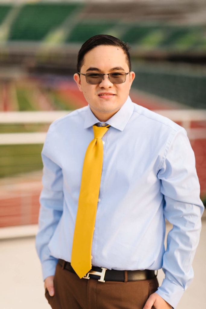 male university of Oregon graduate wearing yellow tie standing in front of fence looking over Hayward Field during graduation photos