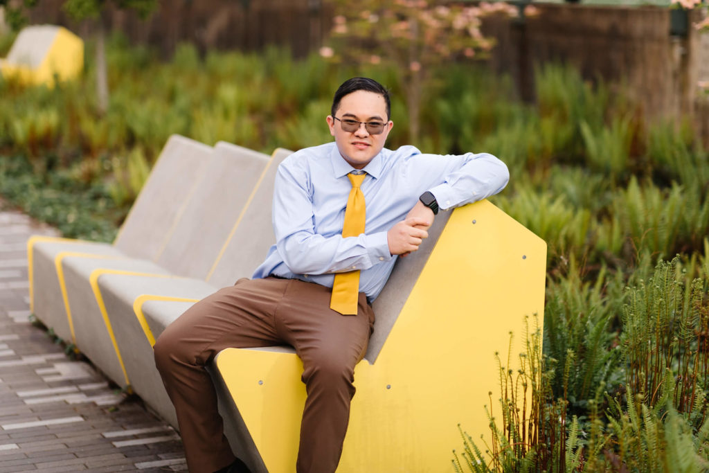 male university of Oregon graduate wearing yellow tie and sitting on yellow bench smiling at camera near Hayward Field during graduation photos