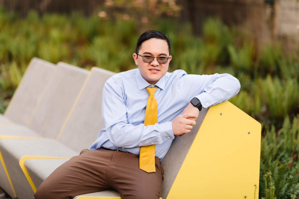 male university of Oregon graduate wearing yellow tie and sitting on yellow bench smiling at camera near Hayward Field during graduation photos