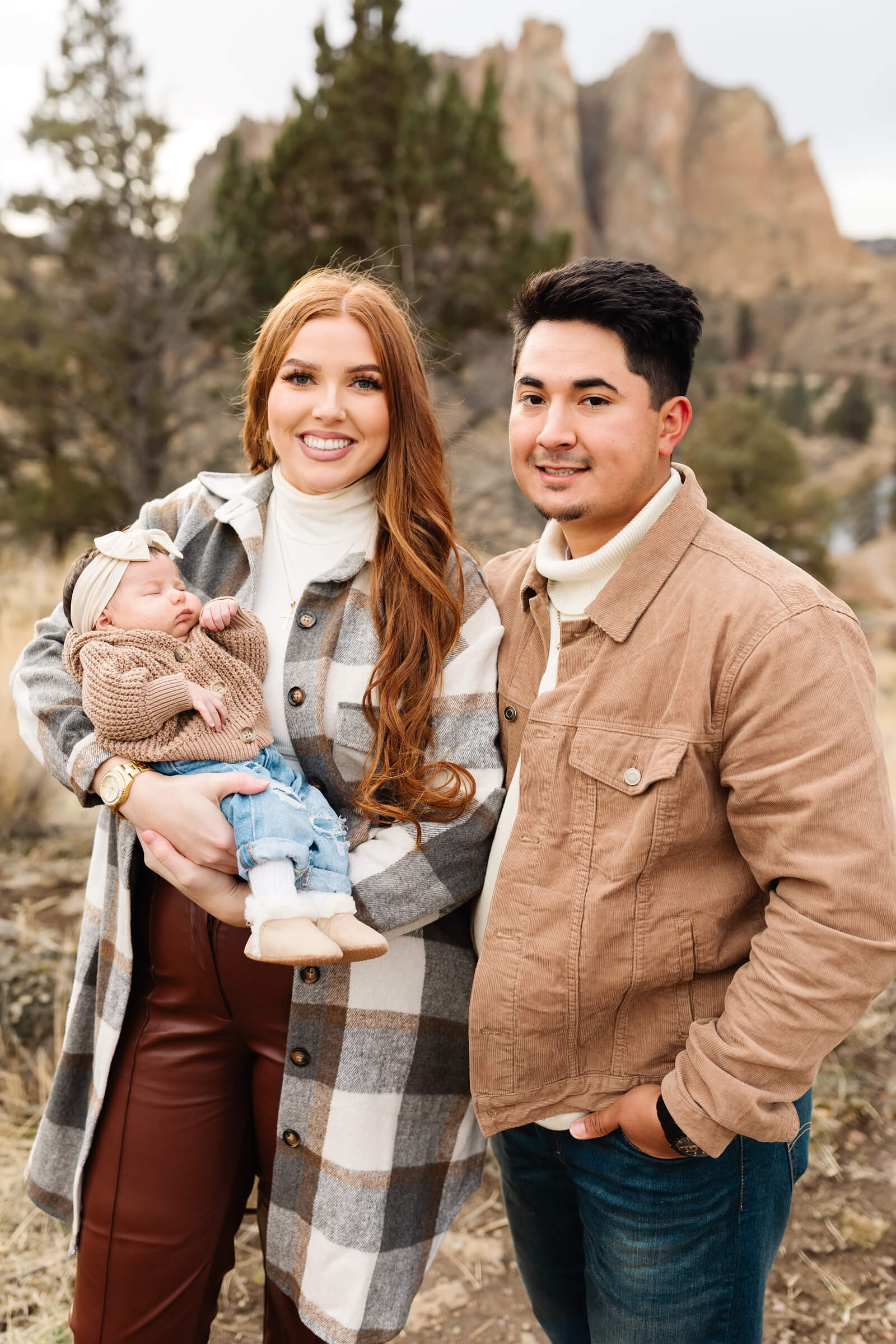 red haired woman in long gray plaid shacket holding newborn baby with cream colored bow on her head during smith rock family photoshoot