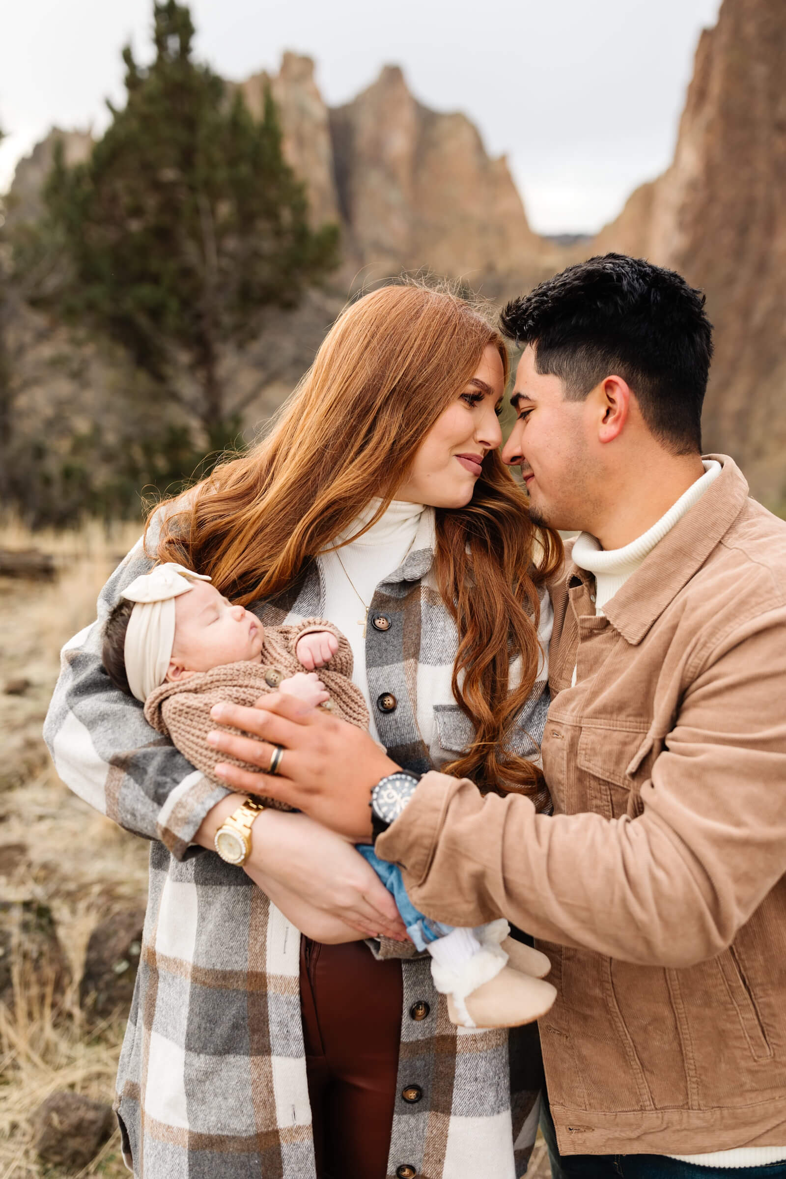 red haired woman in long gray plaid shacket holding newborn baby with cream colored bow on her head nose to nose with husband