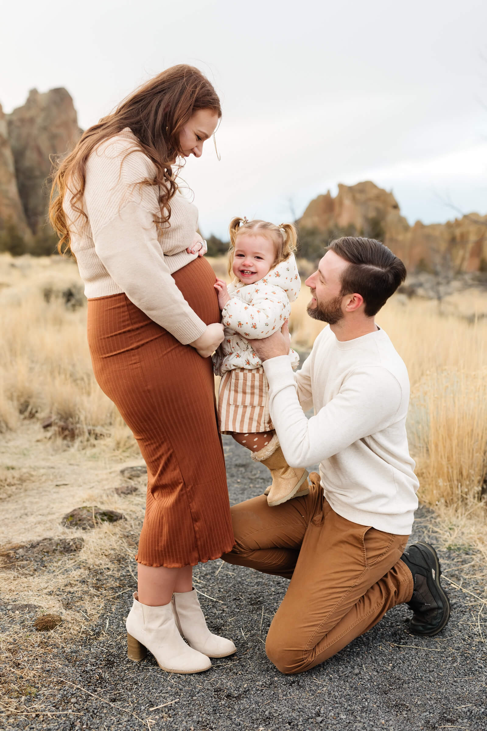 little girl with pigtails smiling at mothers pregnant belly during family photoshoot in November