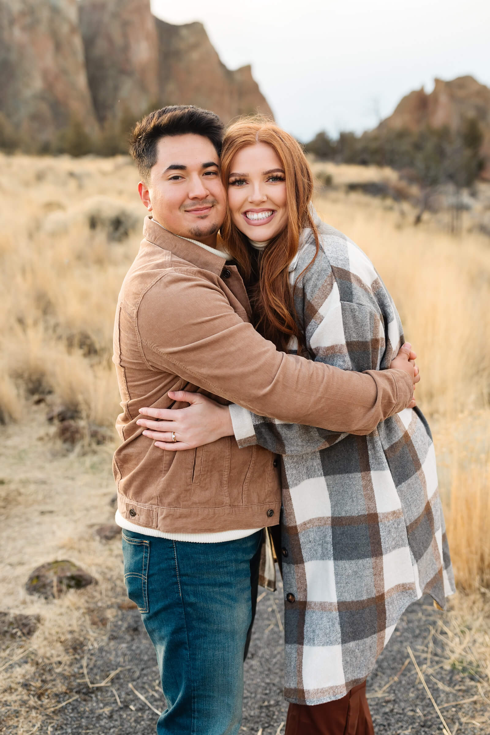 dark haired male in leather jacket and woman with red hair and gray plaid shacket hugging and smiling at smith rock