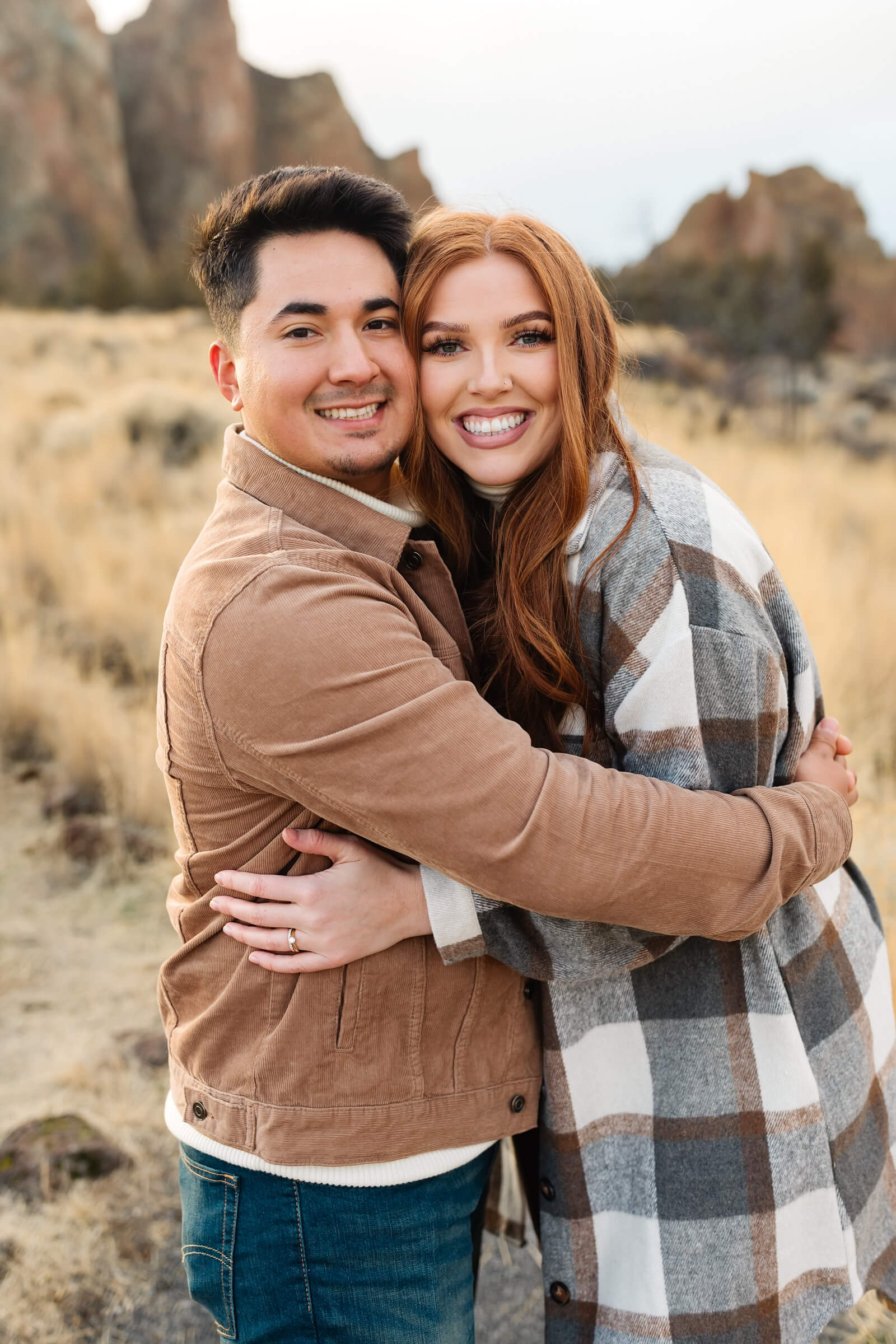 dark haired male in leather jacket and woman with red hair and gray plaid shacket hugging and smiling during smith rock family photoshoot