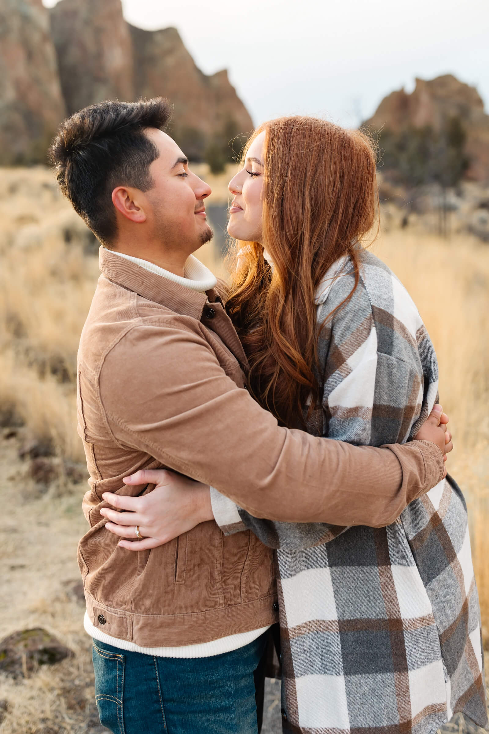 dark haired male in leather jacket and woman with red hair and gray plaid shacket hugging and looking at eachother during smith rock family photoshoot